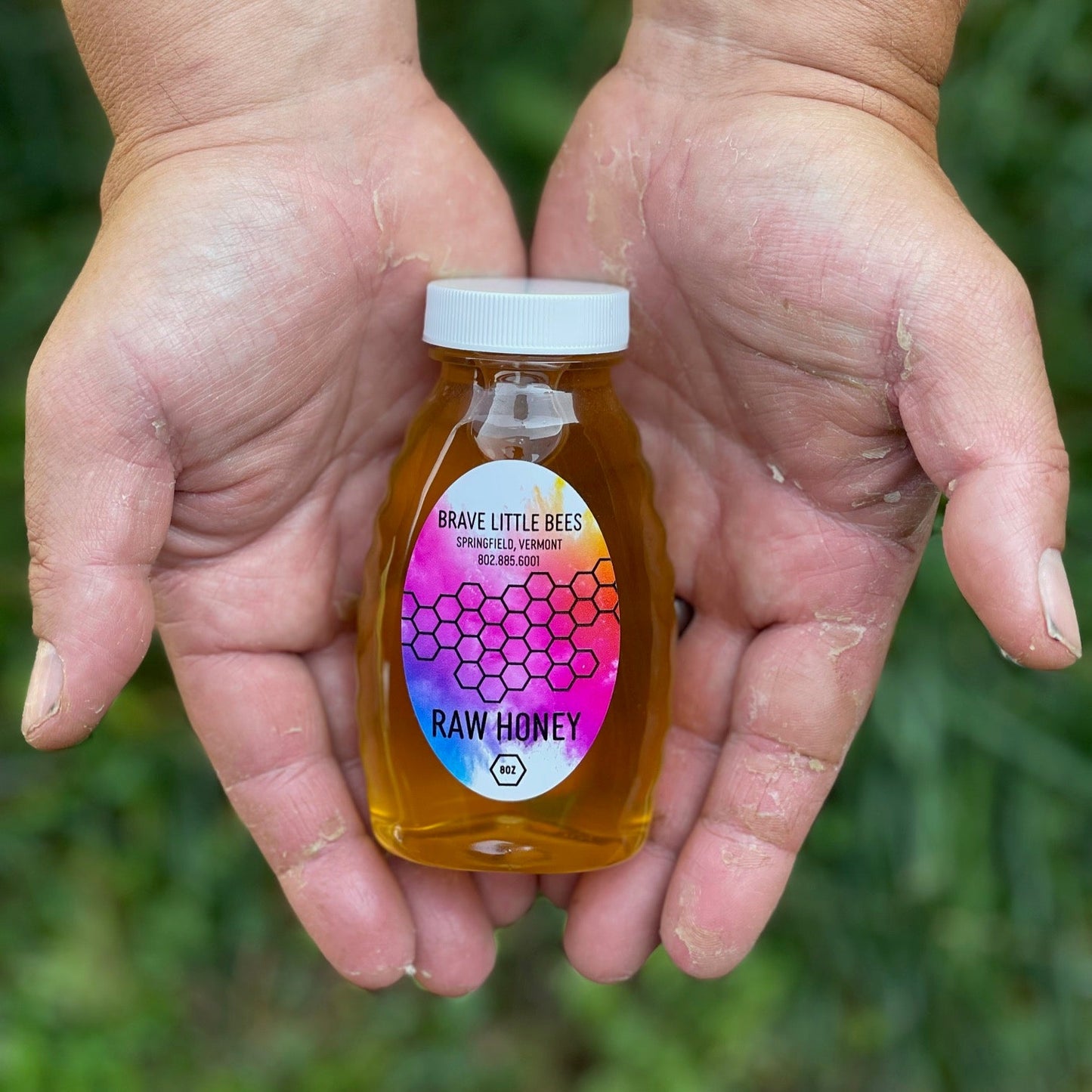 Two hands holding a bottle of raw honey with a colorful label against a natural background.