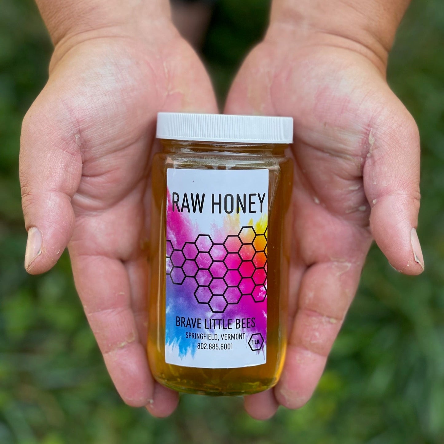 Two hands holding a jar of 'Raw Honey' by 'Brave Little Bees' against a natural background.