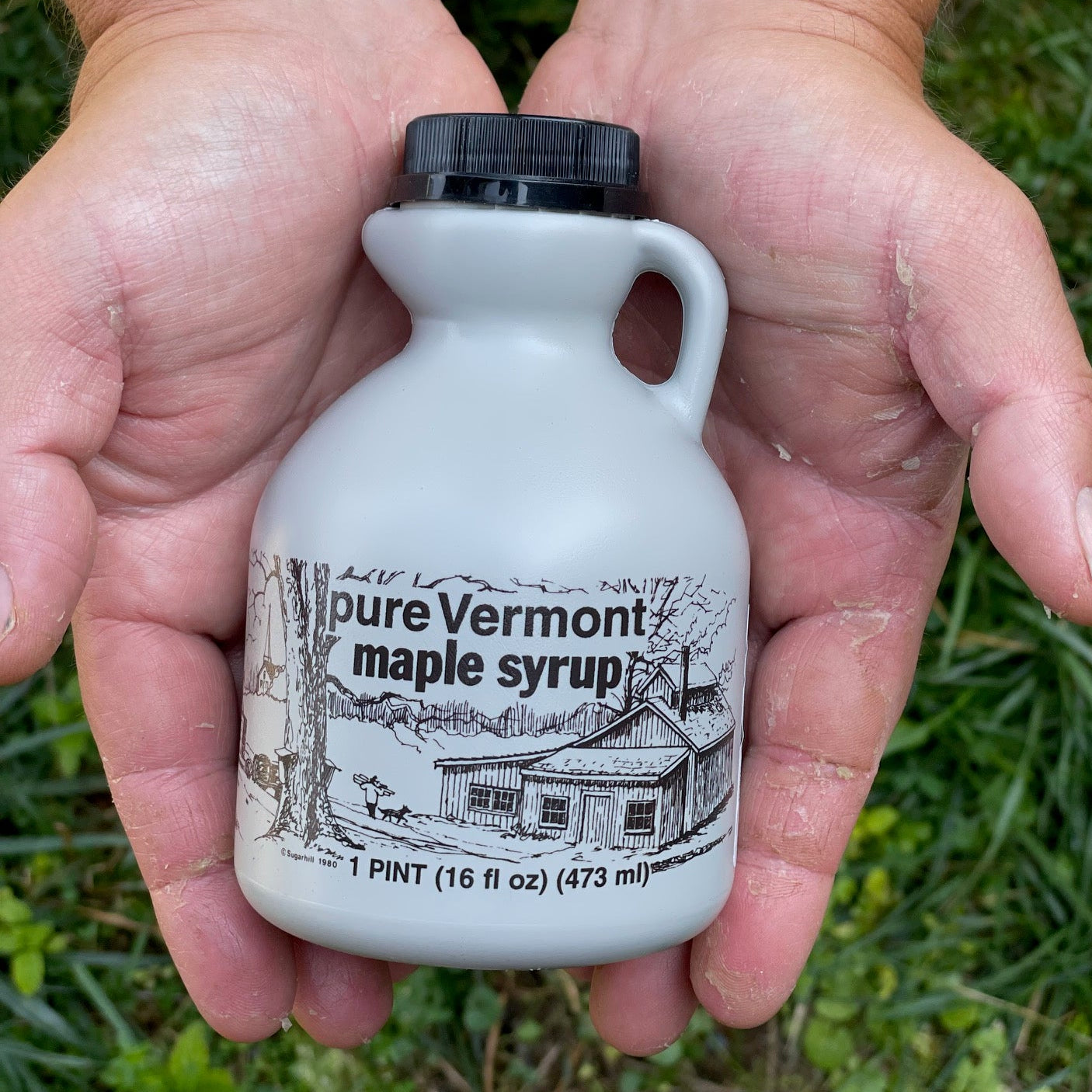 Hand holding a small bottle of pure Vermont maple syrup with a scenic label.